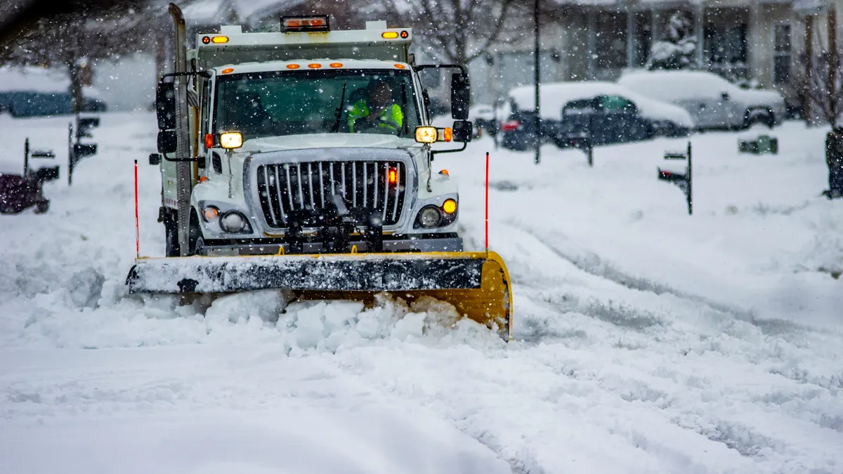 Uno spazzaneve su un grande camion di servizio spinge diversi centimetri di neve verso lo spettatore. Lo spazzaneve si trova su una strada di periferia mentre la neve continua a cadere.